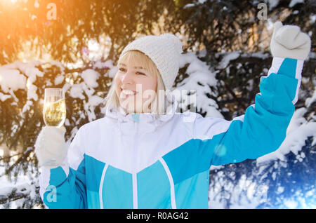 Belle fille blonde avec un verre de champagne en plein air chante et danse sur un fond de sapins enneigés en hiver Banque D'Images