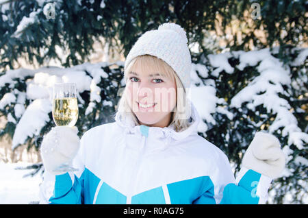 Belle fille blonde avec un verre de champagne en plein air chante et danse sur un fond de sapins enneigés en hiver Banque D'Images