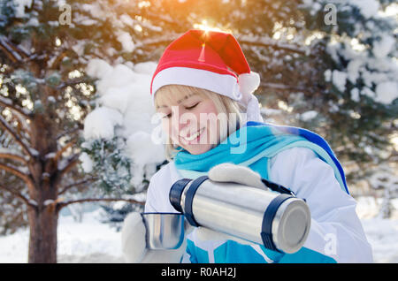 Beautiful smiling blonde girl in a red Christmas hat verse du thé chaud dans une tasse d'un thermos Banque D'Images