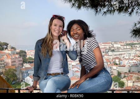 Deux jeunes amis féminins - un Africain, un Européen, assis sur un mur sur une journée d'été avec l'horizon de Lisbonne derrière eux Banque D'Images