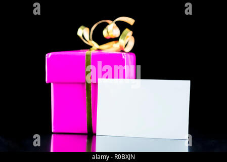 Boîte cadeau rose enveloppé avec archet d'or vierge et carte de vœux. Sur fond noir isolé. Banque D'Images