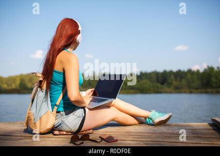 Photo de jeune femme avec un casque à écouteurs avec sac à dos et ordinateur portable en mains assis sur la berge sud du fleuve Banque D'Images