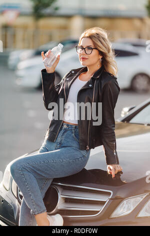 Portrait of young blonde avec bouteille assis sur le capot de voiture noir Banque D'Images