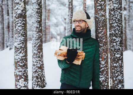 Heureux homme barbu en veste verte, se dresse près de l'arbre, est titulaire du bois de chauffage, l'air songeur de côté, a l'expression joyeuse, se dresse contre la forêt enneigée ba Banque D'Images