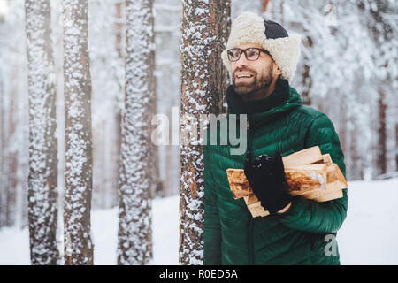 Agréable à heureux homme détient le bois de chauffage, l'air songeur de côté, se dresse près de arbres d'hiver, des rêves à propos de quelque chose d'agréable, jouit de magnifiques wint Banque D'Images