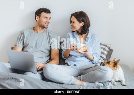 Portrait de lovely couple siéger ensemble au lit, d'utiliser un ordinateur portable, ont un agréable conversation entre eux, faire du shopping en ligne. Jack Russell te Banque D'Images