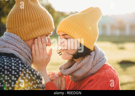 Portrait de femme brune horizontale en chapeau chaud, écharpe et pull, garde les mains sur la joue de petit ami, ont moment romantique, aller à baiser comme datant Banque D'Images