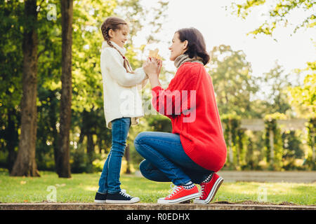 Jeune mère en tricot chaud chandail rouge joue avec sa petite fille dans le parc, lui donne sa feuille, profiter du beau temps d'automne. Maman affectueuse et littl Banque D'Images