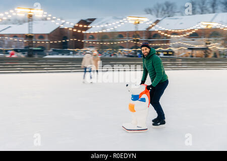 Photo de heureux homme barbu passe du temps en plein air, sur l'air frais, bénéficie d'un hiver glacial, patinage sur l'anneau de glace va, utilise l'aide comme skae peur de tomber n' Banque D'Images