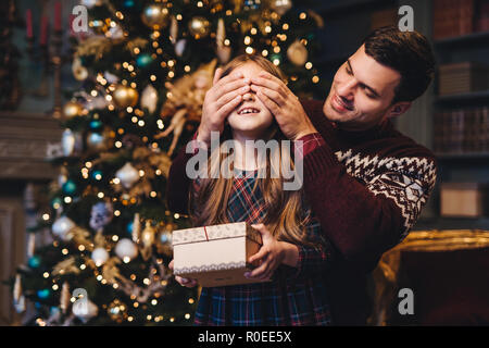 Portrait de jeune père couvre les yeux de sa fille que va faire une surprise pour elle, donne à présent, se tenir ensemble près de l'arbre de Noël. Happy smilin Banque D'Images