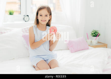 Jolie petite fille est titulaire d'un délicieux donut dans les mains, va prendre le petit-déjeuner, pose sur le lit confortable en chambre blanche, regarde avec appareil photo expres positive Banque D'Images