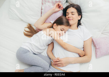 La famille et l'heure du coucher concept. Heureux sleepy mère et son adorable fille embrasser au lit et d'agréables rêves, s'allonger sur les draps blancs, ont rendez-vous Banque D'Images