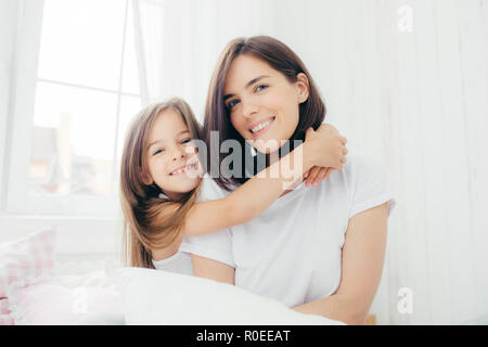 Tourné à l'intérieur de la bonne à la mère brunette avec sourire doux et sa petite fille donne aux hug, profitez de l'atmosphère intérieure, pose contre blanc spacieux Banque D'Images