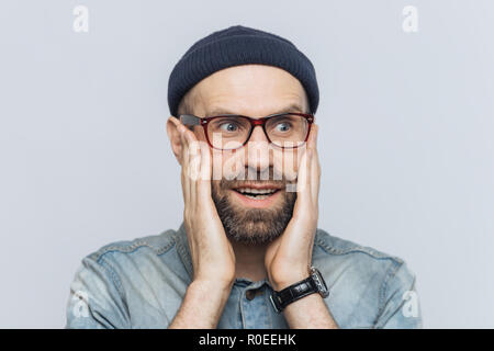 Surpris handsome middle aged man looks en distance avec perplexité, garde les mains sur les joues, porte des lunettes et un chapeau, isolé sur fond blanc. Les Peo Banque D'Images