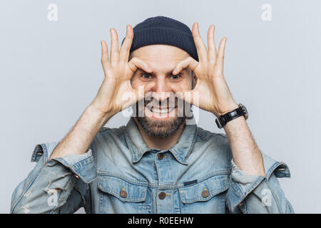 Portrait d'homme barbu hppy lunettes fait avec les doigts, a chaume, porte des jeans vêtements, se dresse contre l'arrière-plan blanc. Homme d'âge moyen drôle foo Banque D'Images