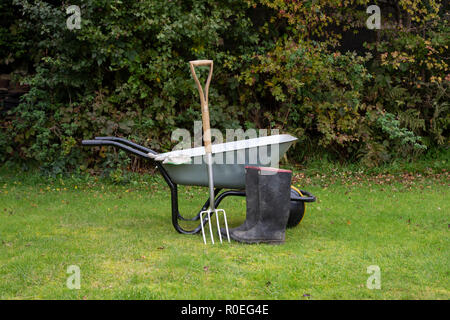 Brouette de jardin, tablier, gants et bottes sur l'herbe contre une haie d'aubépine Banque D'Images