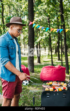 Vue latérale d'african american man in hat et la cuisson des aliments sur le gril dans park Banque D'Images