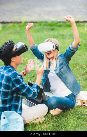 Vue latérale d'étudiants multiculturels dans les casques de réalité virtuelle d'un ordinateur portable assis sur l'herbe verte dans le parc Banque D'Images