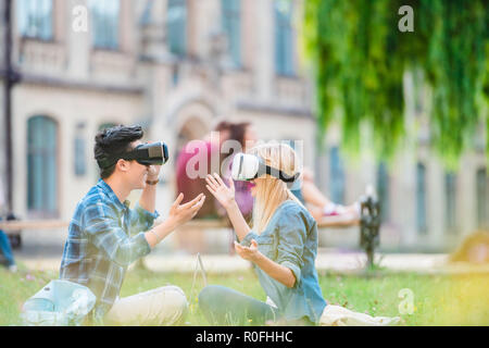 Vue latérale d'étudiants multiculturels dans les casques de réalité virtuelle d'un ordinateur portable assis sur l'herbe verte dans le parc Banque D'Images