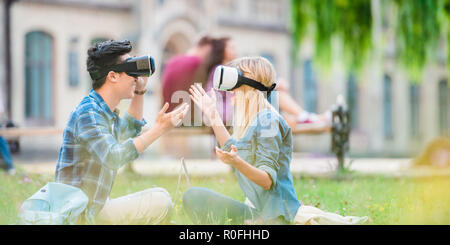 Vue latérale d'étudiants multiculturels dans les casques de réalité virtuelle d'un ordinateur portable assis sur l'herbe verte dans le parc Banque D'Images