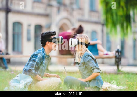 Vue latérale d'étudiants multiculturels dans les casques de réalité virtuelle d'un ordinateur portable assis sur l'herbe verte dans le parc Banque D'Images