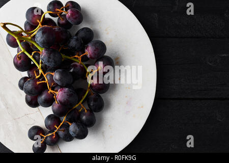 Vue de dessus de raisins noirs frais mûrs sur une assiette de marbre blanc sur noir table en bois avec copie espace. Close up. Banque D'Images