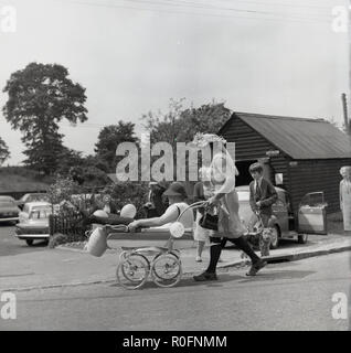 1967, la race, la pram village personnes observant un homme adulte compétiteur dans 'drag'- habillés en vêtements féminins - en poussant son landau et 'bébé' (un homme assis dans le landau), le long d'une route de village, Gerin, Buckinghamshire, Angleterre, Royaume-Uni. Banque D'Images