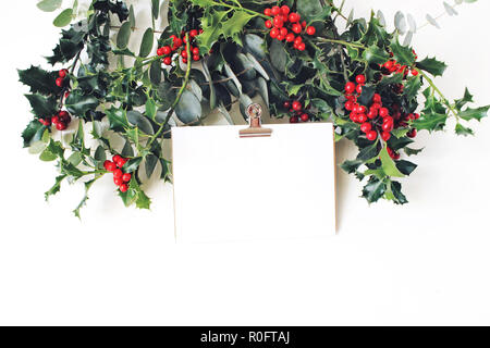 Maquette de Noël scène. Carte de vœux papier doré clip binder, eucalyptus et holly baies rouges, feuilles et branches sur fond de table blanc. Mariage d'hiver. Banque D'Images