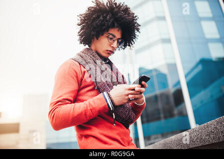 Un beau gars aux cheveux bouclés de race mixte est habillée dans un quartier branché de style jeunesse foulard et sweater écrit un message dans un teleone sur fond de Banque D'Images
