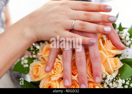 Mariée et le marié les mains avec les anneaux de mariage contre l'arrière-plan de bouquet de fleurs. Déclaration d'amour, au printemps. Carte de mariage, la Saint Valentin message d'accueil. Les anneaux de mariage. Détails de jour de mariage Banque D'Images