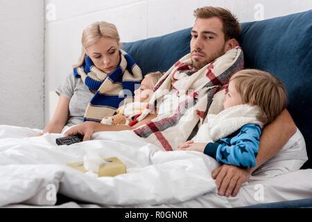 Les jeunes malades dans la famille des écharpes au lit avec la télécommande du téléviseur Banque D'Images