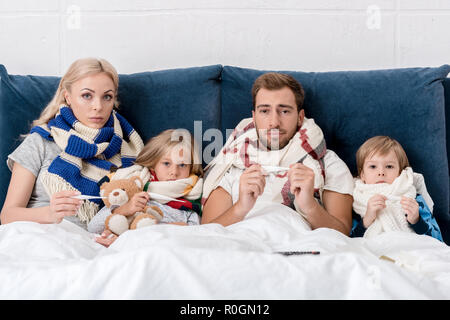 Les jeunes malades dans la famille des écharpes holding electric thermomètres et looking at camera while Lying in Bed Banque D'Images