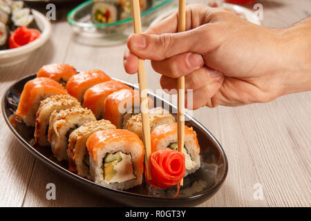 Homme main avec deux baguettes holding gingembre mariné sous forme de rose rouge et différents rouleaux de sushi avec du riz, légumes, fruits de mer sur plaque en céramique Banque D'Images