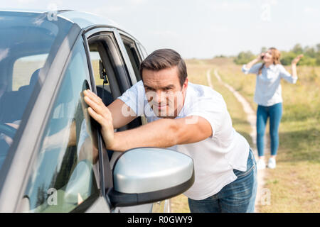 Man pushing broken voiture pendant que sa femme à parler par téléphone dans le champ au cours de déplacement en voiture Banque D'Images