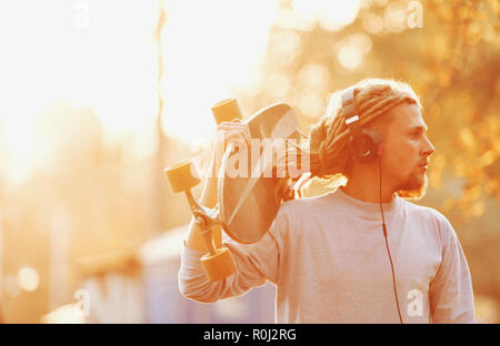 Patineur hippie homme barbu blond avec des dreads portant des écouteurs marche dans la forêt et à la recherche sur le coucher du soleil, vue extérieure Banque D'Images