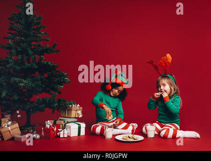 Deux petites filles vêtues de vêtements thème Noël assis à côté d'un arbre de Noël. Enfants heureux, assis près d'un arbre de Noël profitant des cookies avec Banque D'Images