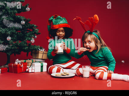 Deux enfants assis à côté d'un arbre de Noël de manger des cookies avec du lait. Petites filles dans fantaisie vêtements de Noël assis à côté d'un arbre de Noël avec des cadeaux. Banque D'Images