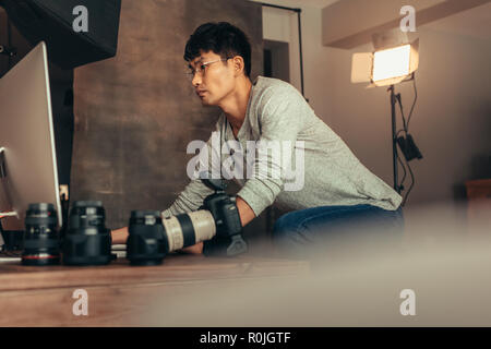 La configuration de son ordinateur, photographe pour le tournage en studio. L'homme travaillant sur ordinateur avec caméra et objectif sur table. Banque D'Images