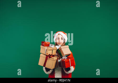 Robe fille au père Noël tenant une pile de boîtes cadeaux. Kid habillé comme santa holding gifts debout sur un fond vert. Banque D'Images