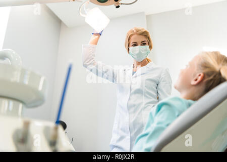 Vue de dessous de jeune femme dentiste avec lampe au-dessus de celui du petit enfant en fauteuil Banque D'Images