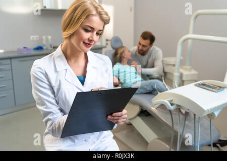 Femme dentiste écrit dans le presse-papiers alors que petit enfant et son père assis sur l'arrière-plan Banque D'Images