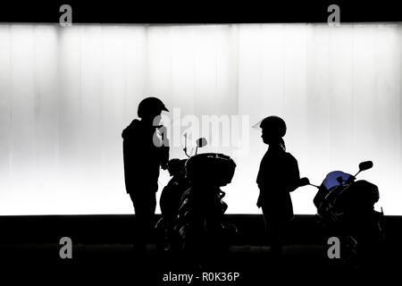 La silhouette floue de l'homme et de la femme de mettre un casque et la préparation de s'asseoir sur une moto en face de bâtiment moderne dans la nuit en noir et blanc Banque D'Images
