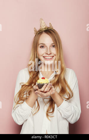 Jeune femme, d'une couronne, tenant un petit gâteau d'anniversaire avec une bougie et cerise sur le dessus sur fond rose Banque D'Images