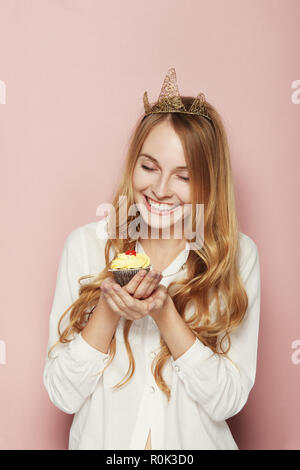 Jeune femme, d'une couronne, tenant un petit gâteau d'anniversaire avec une bougie et cerise sur le dessus sur fond rose Banque D'Images