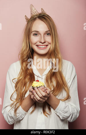 Jeune femme, d'une couronne, tenant un petit gâteau d'anniversaire avec une bougie et cerise sur le dessus sur fond rose Banque D'Images