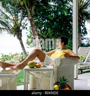 Jeune femme se détendre dans un fauteuil en rotin blanc, terrasse, jardin tropical, Guadeloupe, French West Indies, Banque D'Images