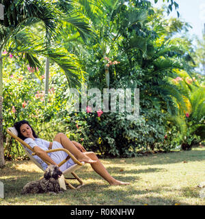 Jeune femme assise dans une chaise et caressant son chien, d'un jardin tropical, Guadeloupe, French West Indies, Banque D'Images