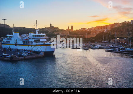 Apprécier le coucher du soleil sur la mer à partir de 13/15 avec un V iew sur les collines de la ville, Mgarr Harbour et ferry, amarré à la rive, Gozo, Malte. Banque D'Images