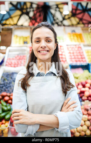 Jeune femme confiante positive en épicerie apron crossing arms sur la poitrine et à la caméra tout en se tenant à l'encontre de la nourriture fraîche caler au farmers market Banque D'Images