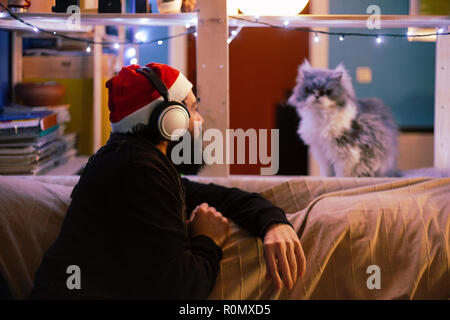 Jeune homme avec un casque et christmas hat en regardant son chat sur un canapé avec lumières décorations Banque D'Images